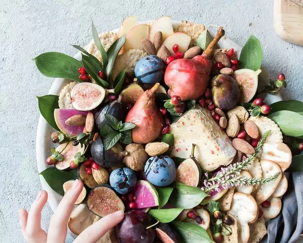 Overhead view of a vibrant breakfast platter with figs, pears, plums, cheese, crackers, nuts, and pomegranates, with a hand r