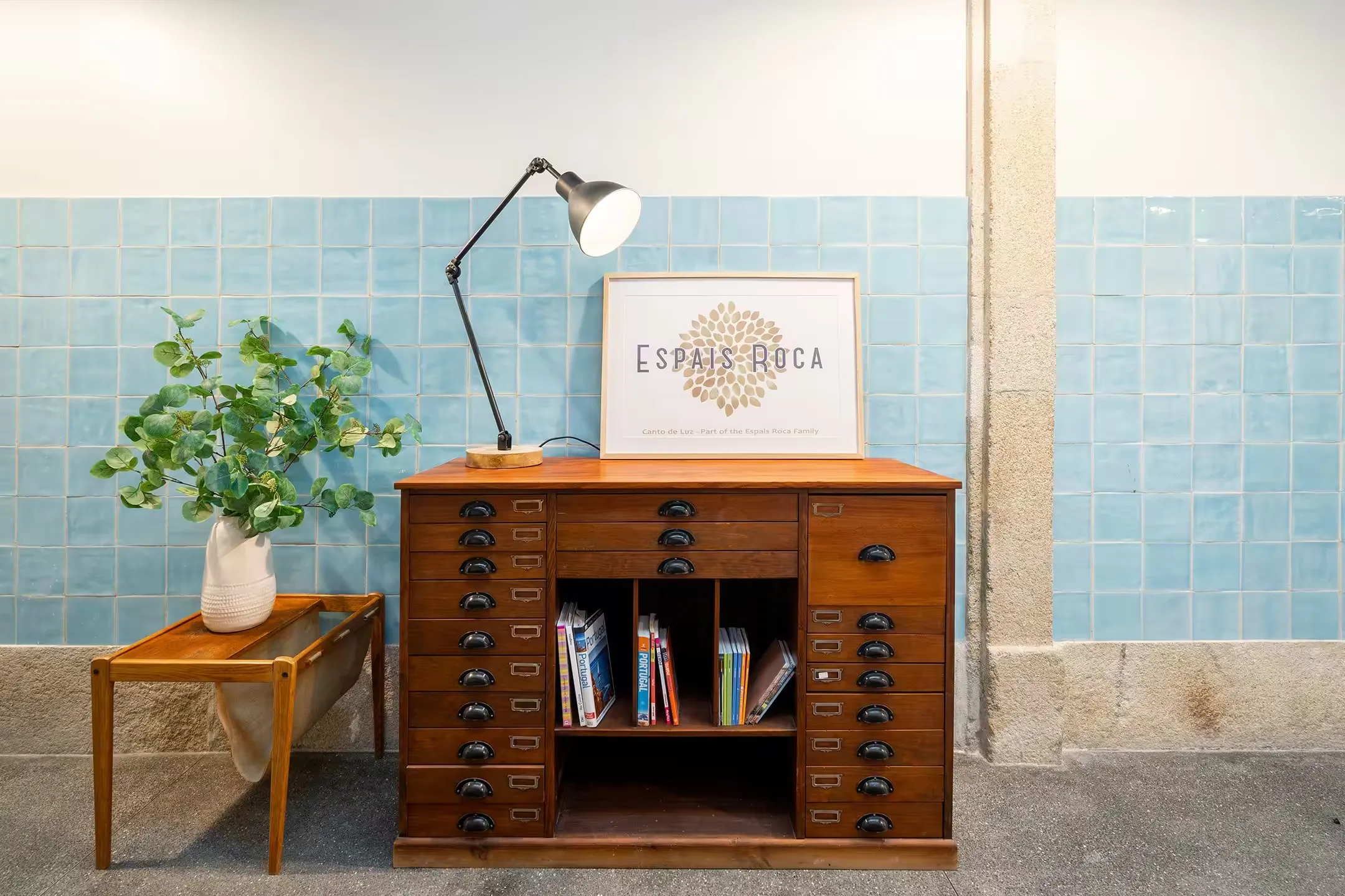 Vintage wooden chest of drawers with black handles, a desk lamp, and a framed 'Espais Roca' print against a blue tiled wall.