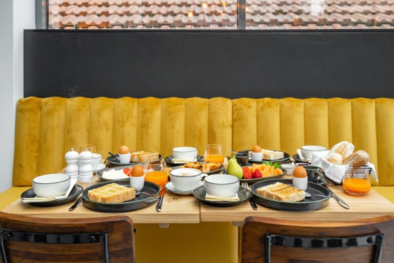A breakfast spread with toast, boiled eggs, fruit, and orange juice on a wooden table with a yellow upholstered bench.