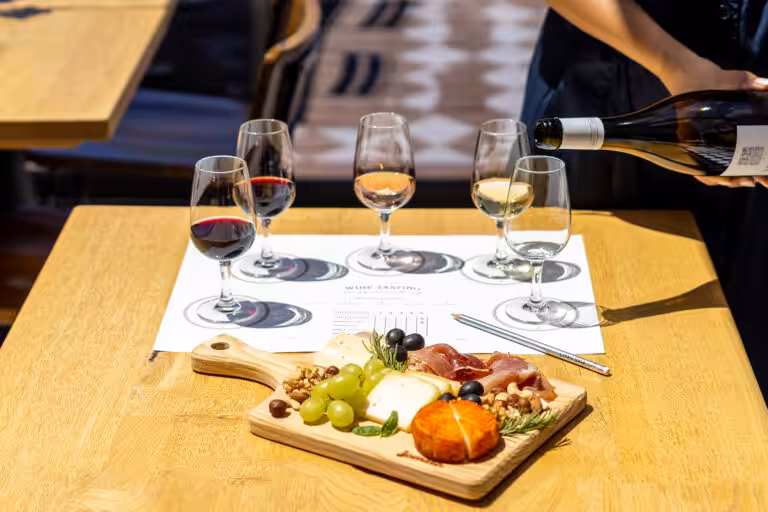 A person pouring white wine into a glass during a wine tasting with a charcuterie board on a wooden table at 14 Acres.