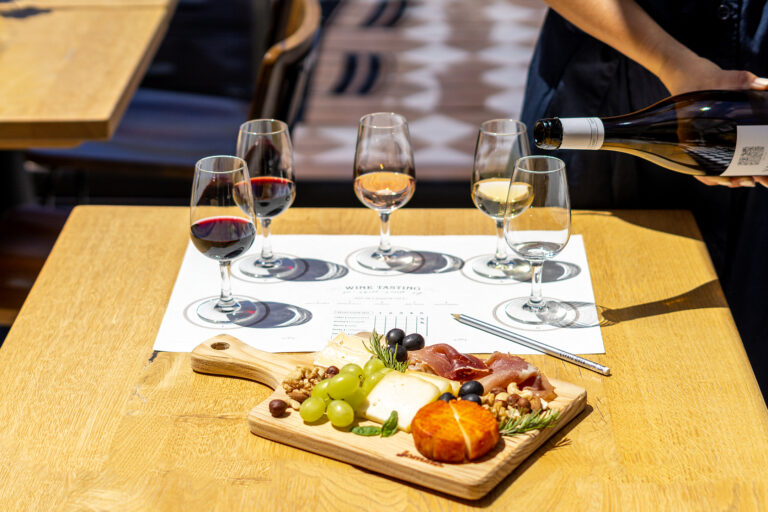 A person pouring white wine into a glass during a wine tasting with a charcuterie board on a wooden table at 14 Acres.