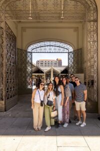 A group of seven smiling friends, three men and four women, stand together in front of an ornate arched entryway, with a bust