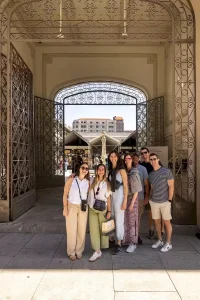 A group of seven smiling friends, three men and four women, stand together in front of an ornate arched gateway. Beyond them,