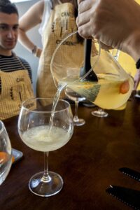 Close-up of a hand pouring a refreshing fruit-infused drink from a pitcher into a wine glass, with people in aprons in the ba