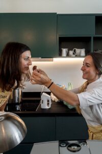 Two women in a modern kitchen, one in a chef's jacket offering a small chocolate treat to the other. Dark green cabinets, sle