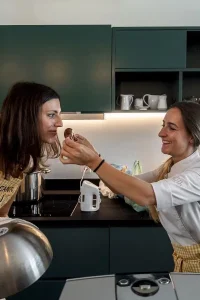 Two women in a modern kitchen, one in a chef's jacket feeding a chocolate truffle to the other, enjoying a cooking class at 1