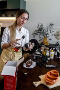 Woman in chef's jacket and yellow apron pouring Alianca brandy into a small black cup, preparing food in a kitchen.