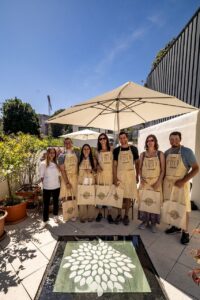 Six people, including a chef, pose with cooking aprons and tote bags on a sunny outdoor patio, ready for a culinary experienc