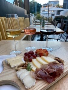 A wooden board with various cheeses, prosciutto, grapes, and nuts, paired with three glasses of white, rosé, and red wine, on