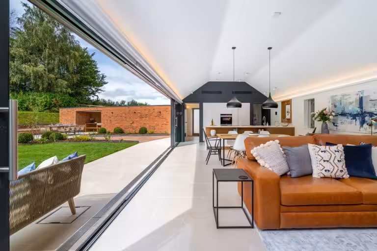 Spacious open-plan living area with a tan leather sofa, modern kitchen, and dining table, opening onto a patio and garden at