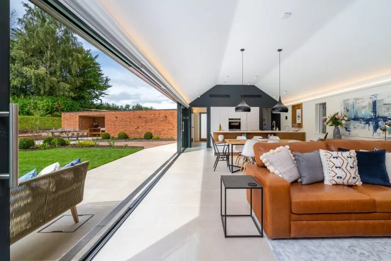 Spacious open-plan living area with a tan leather sofa, modern kitchen, and dining table, opening onto a patio and garden at