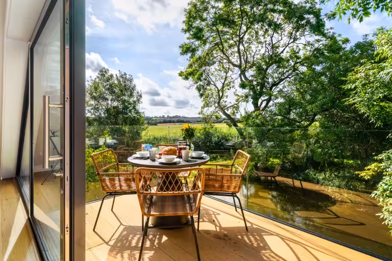 Outdoor dining table set for breakfast on a sunny balcony overlooking a pond and green fields at 14 Acres, Clipston.