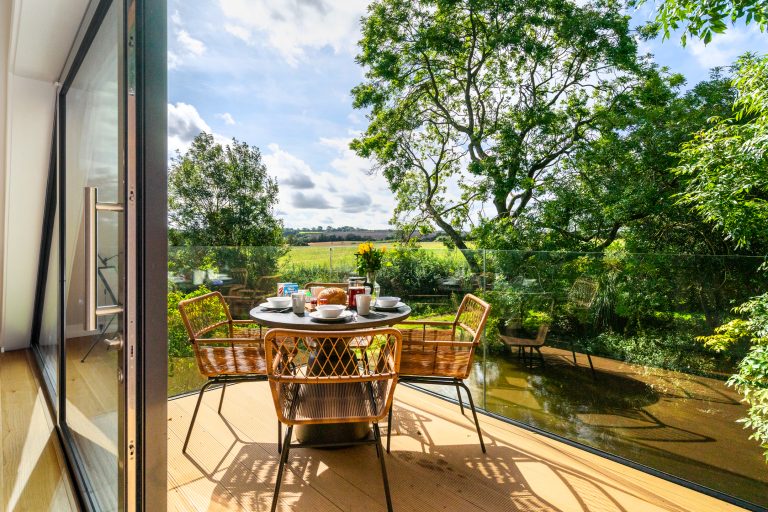 Outdoor dining table set for breakfast on a sunny balcony overlooking a pond and green fields at 14 Acres, Clipston.