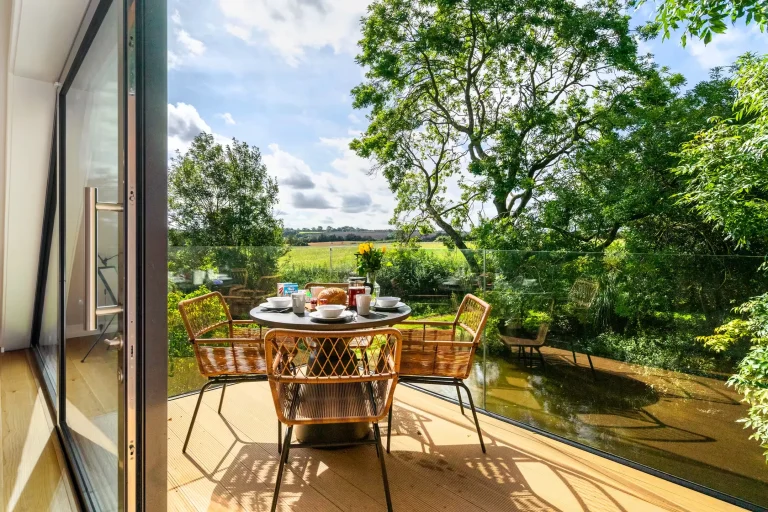 Outdoor dining table set for breakfast on a sunny balcony overlooking a pond and green fields at 14 Acres, Clipston.