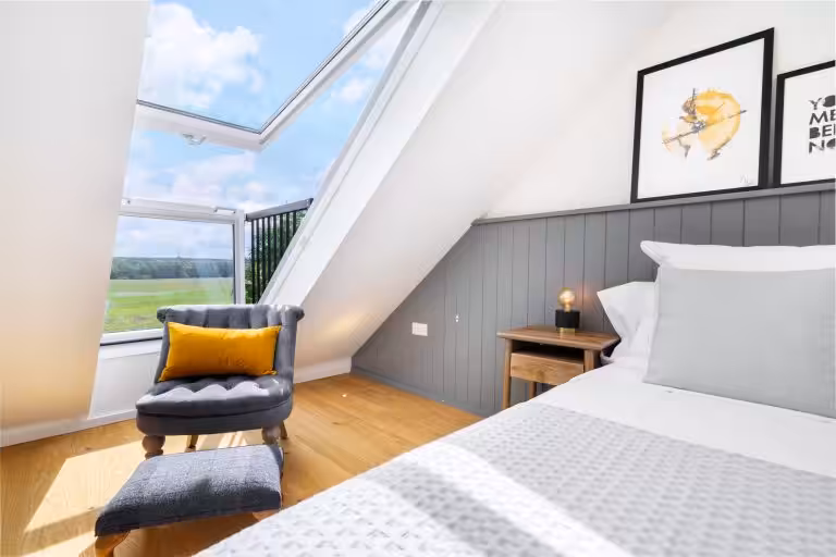Modern bedroom with a grey armchair, yellow cushion, and open skylight overlooking countryside fields at 14 Acres The Loft.