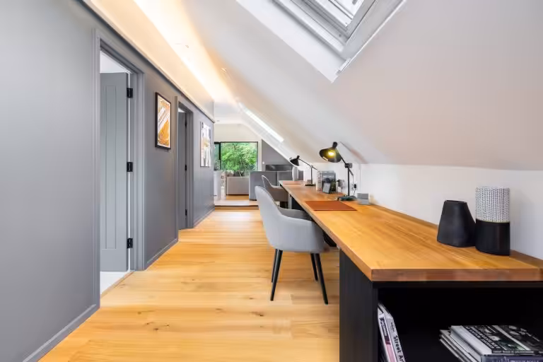 Modern attic workspace with two grey chairs at a long wooden desk, under a skylight, with a living area visible beyond.