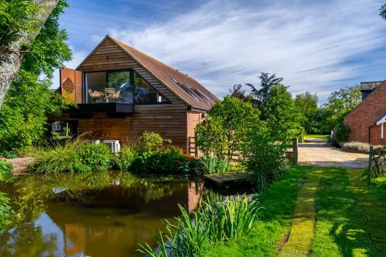 The Barn luxury retreat at 14 Acres, with a modern glass balcony overlooking a tranquil pond and lush gardens under a blue sk