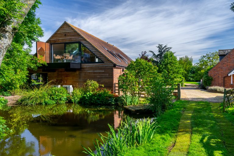 The Barn luxury retreat at 14 Acres, with a modern glass balcony overlooking a tranquil pond and lush gardens under a blue sk