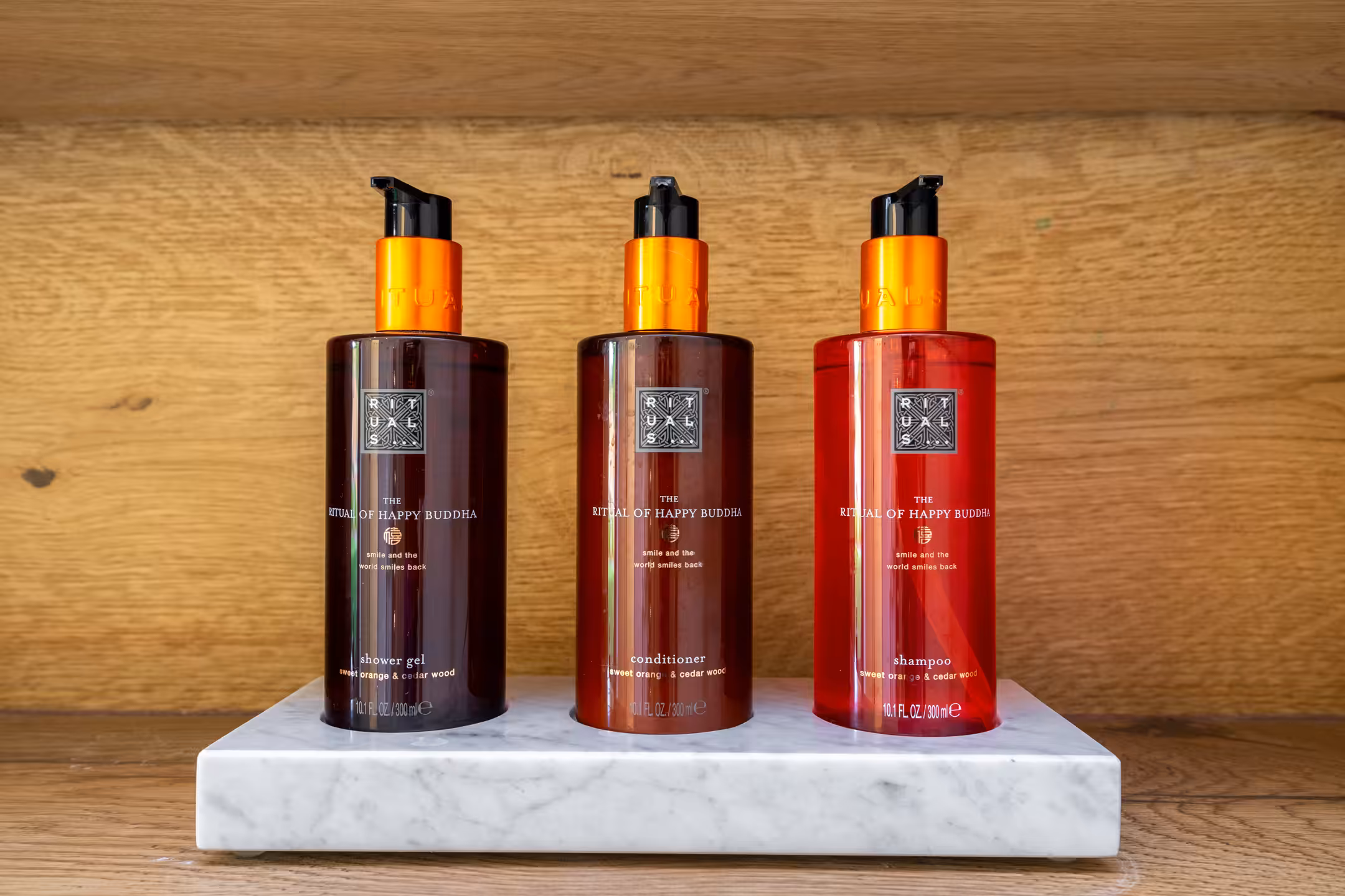 Three Rituals 'The Ritual of Happy Buddha' shower gel, conditioner, and shampoo bottles on a marble tray against a wooden bac