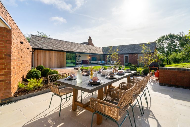 Outdoor dining table set for a meal on a sunny patio at The Barn, 14 Acres, Clipston, with modern architecture and garden.