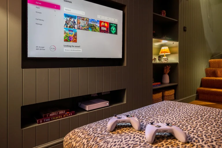 Xbox console and controllers on a leopard print ottoman, with a large TV displaying game options in a luxury lodge at 14 Acre