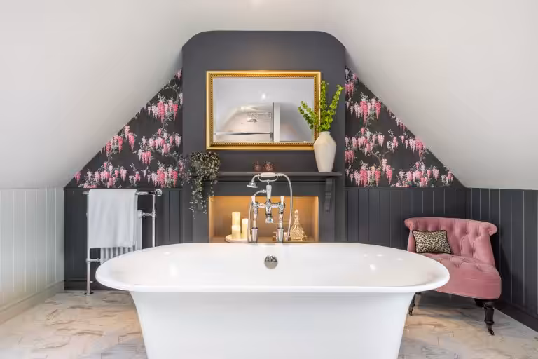 A luxurious white roll-top bathtub in a bathroom with dark panelled walls and pink floral wallpaper, featuring a gold mirror