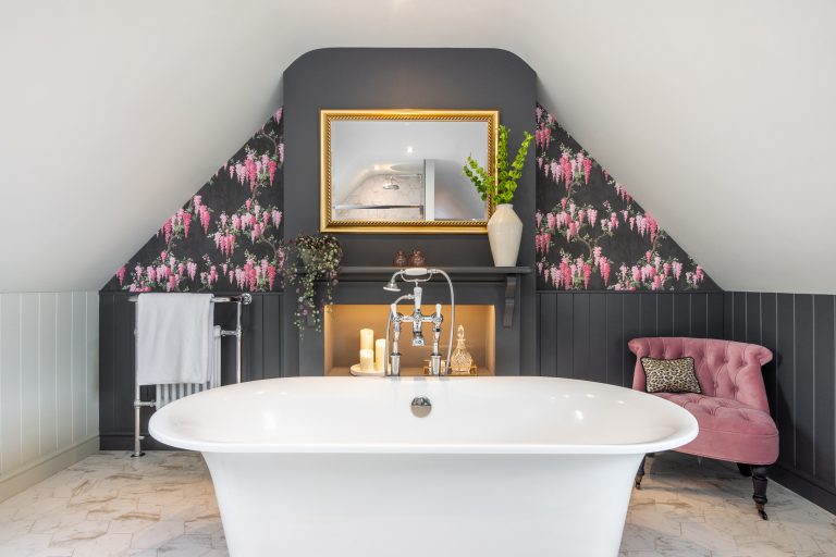 A luxurious white roll-top bathtub in a bathroom with dark panelled walls and pink floral wallpaper, featuring a gold mirror