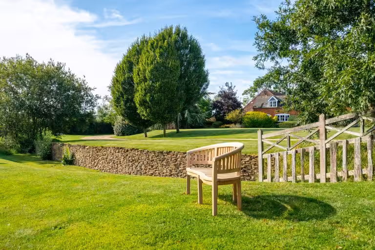 Curved wooden garden bench on a lush green lawn with a dry stone wall, trees, and a traditional brick house in the background