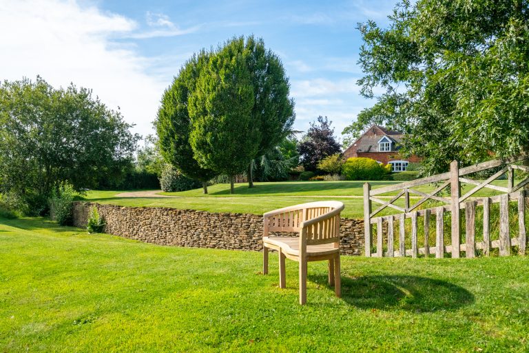 Curved wooden garden bench on a lush green lawn with a dry stone wall, trees, and a traditional brick house in the background