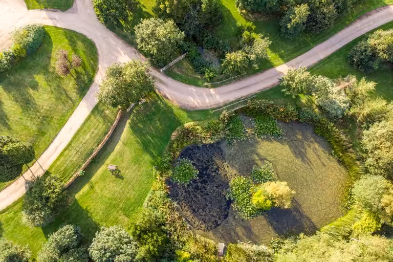 Aerial view of 14 Acres estate with winding gravel paths, lush green lawns, mature trees, and a tranquil pond with lily pads.