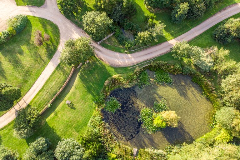 Aerial view of 14 Acres estate with winding gravel paths, lush green lawns, mature trees, and a tranquil pond with lily pads.