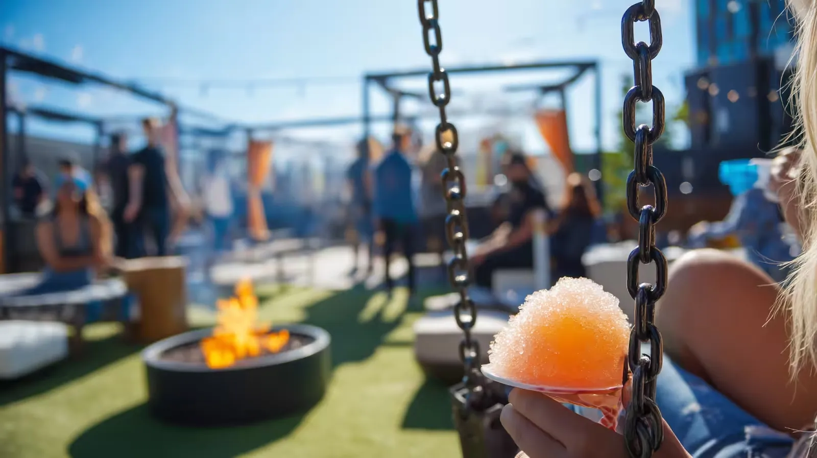 Festival-goer holding a peach slushie on a swing, with a fire pit and crowds behind