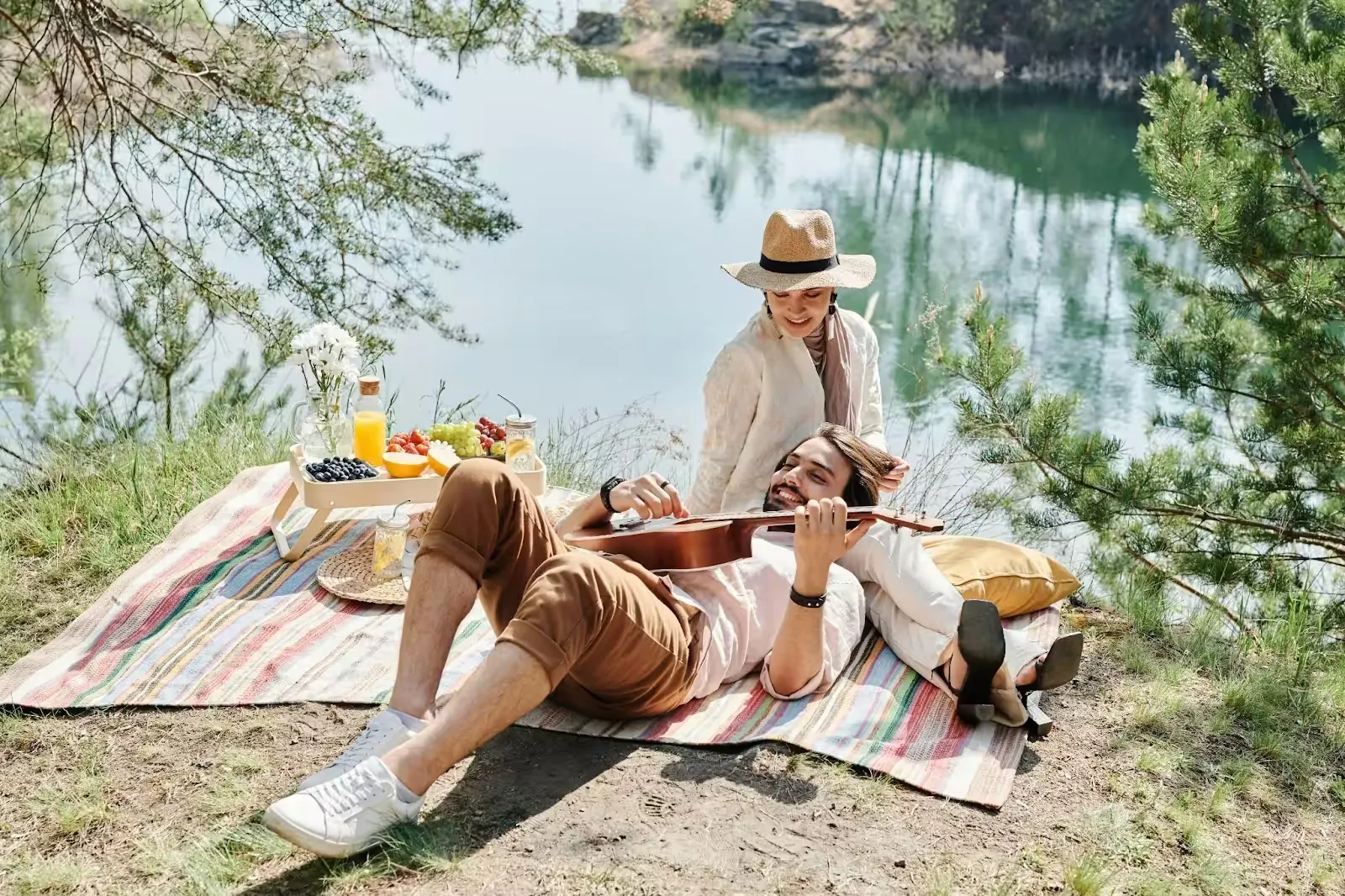 Couple picnicking on a striped blanket beside a lake, one playing a guitar